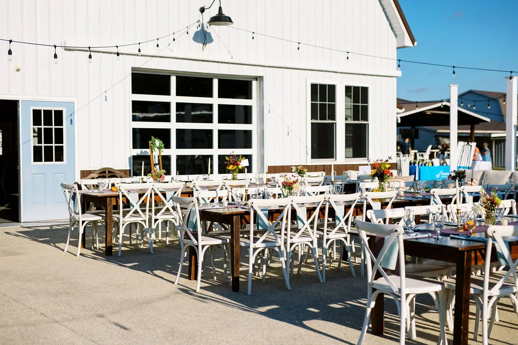 farmhouse tables and cross-back chairs set up outside of a white barn for open air dining.