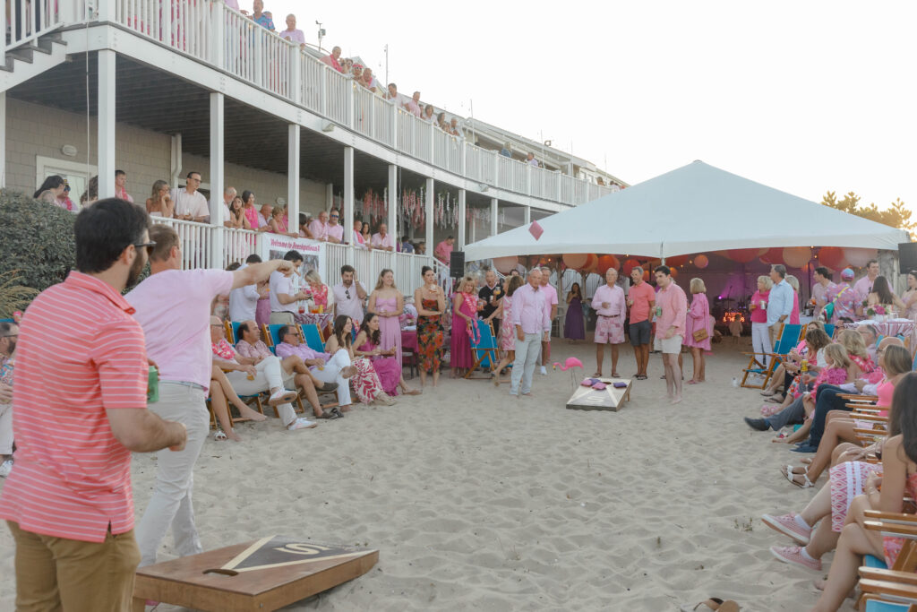 people playing cornhole on the sand at a pink themed party with tent in background